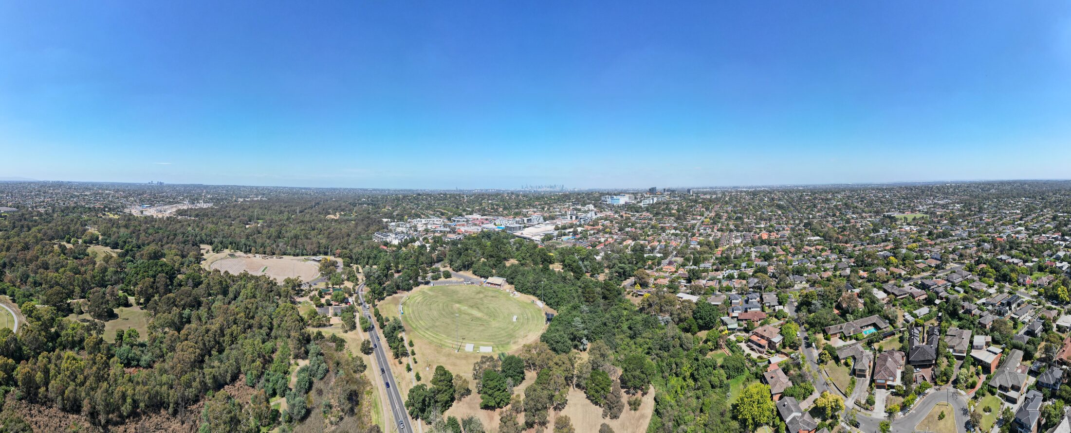 Aerial panorama of Heidelberg showing parkland, mature tree canopy, suburban streets and the Melbourne skyline in the distance